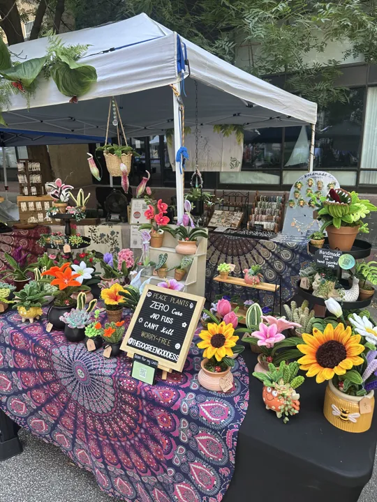 The Go Green Goddess tent at an outdoor market, a Peace Plants sign among tables of wire plants and flowers